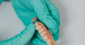 Gloved hands holding a removable partial denture with a pink acrylic base and metal clasps for missing teeth