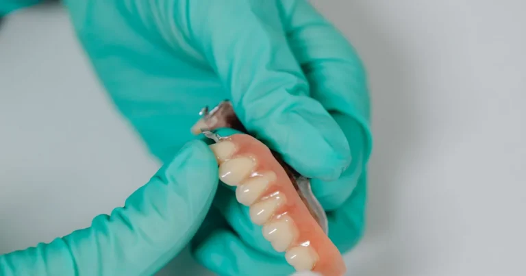 Gloved hands holding a removable partial denture with a pink acrylic base and metal clasps for missing teeth